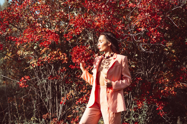 Young beautiful brunette girl with good makeup on her face with expressive features and figure. walks in the fall among beautiful red trees, she is wearing a stylish pink suit and she is a little naked - Foto, Imagem
