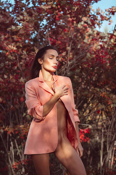 Young beautiful brunette girl with good makeup on her face with expressive features and figure. walks in the fall among beautiful red trees, she is wearing a stylish pink suit and she is a little naked - Foto, Imagen