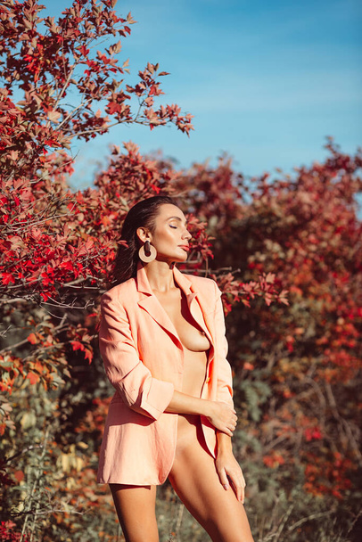 Young beautiful brunette girl with good makeup on her face with expressive features and figure. walks in the fall among beautiful red trees, she is wearing a stylish pink suit and she is a little naked - 写真・画像