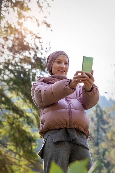 Beyaz ceketli ve yün takım elbiseli genç bir kadın yeşil çam ormanlarında yürürken arkadaşlarıyla ya da ailesiyle cep telefonuyla sohbet ediyor. - Fotoğraf, Görsel
