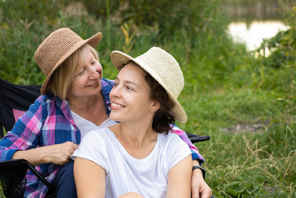 夏のわら帽子のLGBTレズビアンカップルは、週末を一緒に過ごす、湖や川の近くのキャンプ場で椅子に座って笑っています。愛の瞬間幸福の概念 - 写真・画像