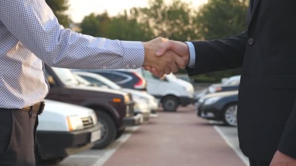 Dois homens de negócios apertando as mãos um do outro com carros no estacionamento no fundo. Aperto de mão entre gerente ou revendedor e cliente. Agitação de braços masculinos lá fora. Fechar câmera lenta - Filmagem, Vídeo