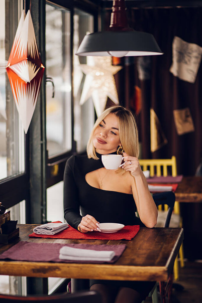 Mujer de negocios sonriente en un restaurante sosteniendo una taza de té y mirando a la cámara, sentada cerca de la ventana con un vestido negro - Foto, imagen