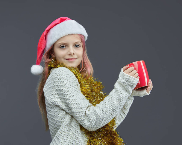 young girl in a Santa hat holds a pink Cup. photo session in the Studio on a gray background - Foto, immagini