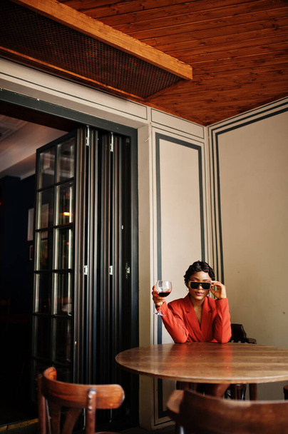 Retrato de mulher afro-americana, penteado retrô com óculos, use jaqueta laranja posando no restaurante com copo de vinho. - Foto, Imagem