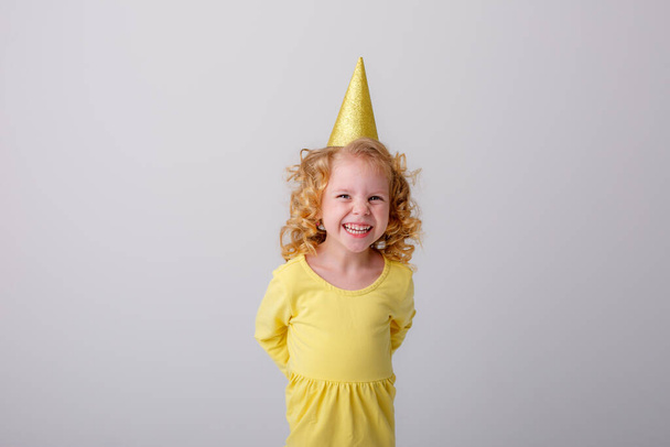 niña en un vestido amarillo en sombrero de cumpleaños posando sobre fondo gris  - Foto, imagen