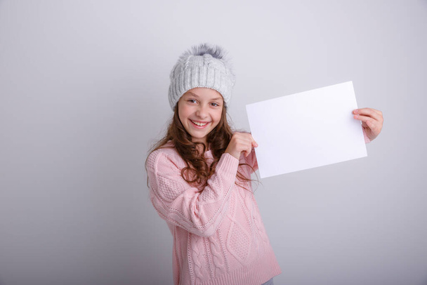 mooi meisje in een witte trui met een blanco vel papier op een grijze achtergrond - Foto, afbeelding