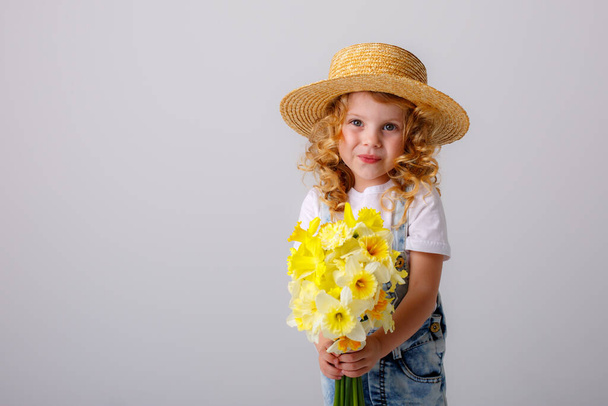 portret van emotionele baby meisje in jeans overall en stro hoed met boeket van narcis bloemen tegen grijs - Foto, afbeelding