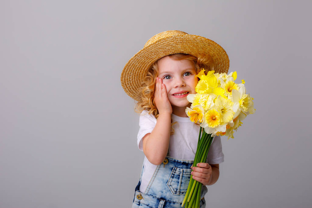 portrait of emotional baby girl in jeans overall and straw hat holding bouquet of daffodil flowers against gray - 写真・画像