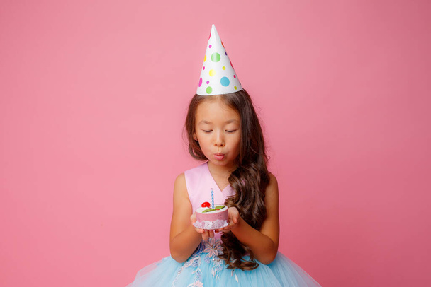 A young Asian girl blows out a candle on a pink background at a birthday party - Foto, Imagem