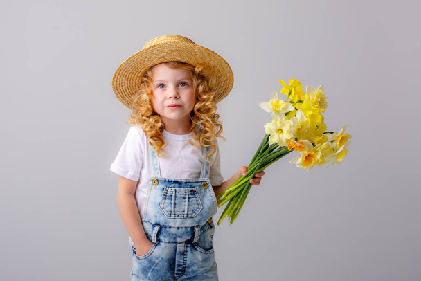 portrait of emotional baby girl in jeans overall and straw hat holding bouquet of daffodil flowers against gray - Photo, Image