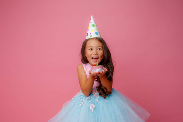 Ein junges asiatisches Mädchen hält auf einer Geburtstagsparty eine Torte mit einer Kerze auf rosa Hintergrund - Foto, Bild