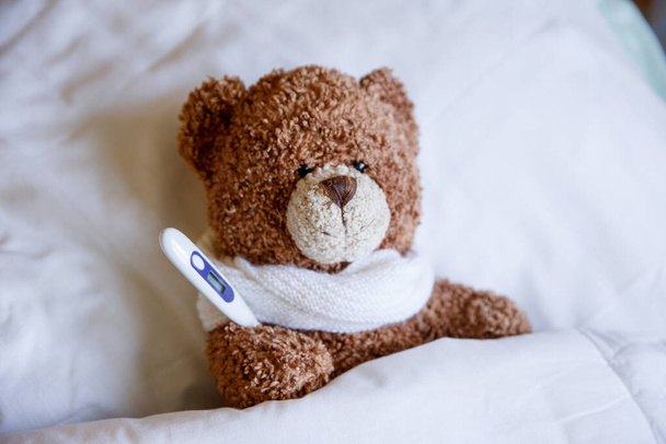 un oso de peluche enfermo está acostado en la cama con un termómetro - Foto, imagen