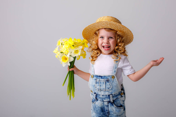 portrait of emotional baby girl in jeans overall and straw hat holding bouquet of daffodil flowers against gray - Foto, afbeelding