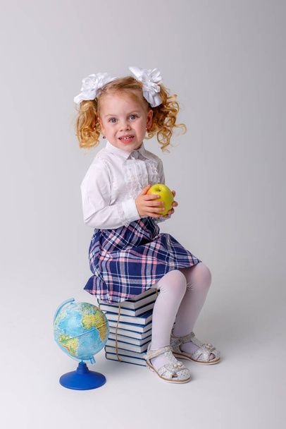 Μια μικρή σγουρομάλλα μαθήτρια με σχολική στολή που κρατάει ένα μήλο στα χέρια της, κάθεται σε μια στοίβα από βιβλία - Φωτογραφία, εικόνα