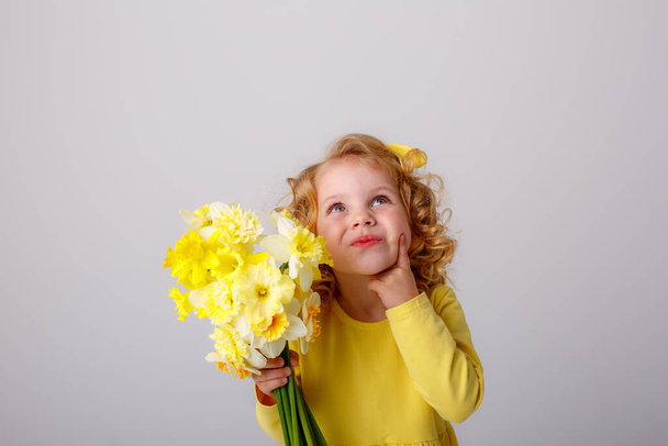 ein kleines gelocktes Mädchen in einem gelben Kleid mit einem Strauß Frühlingsblumen   - Foto, Bild