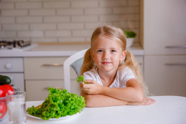 blonde baby girl eating vegetables in the kitchen - Φωτογραφία, εικόνα
