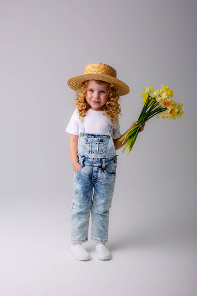 portrait of emotional baby girl in jeans overall and straw hat holding bouquet of daffodil flowers against gray - Foto, afbeelding