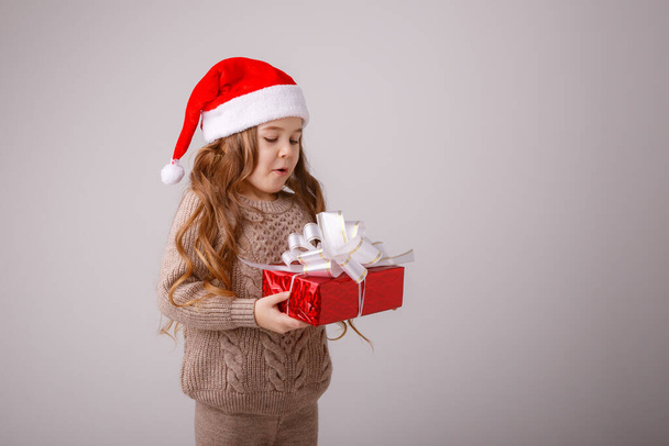 Küçük bir kız, Noel Baba şapkalı. Elinde gri bir hediye kutusu var.  - Fotoğraf, Görsel