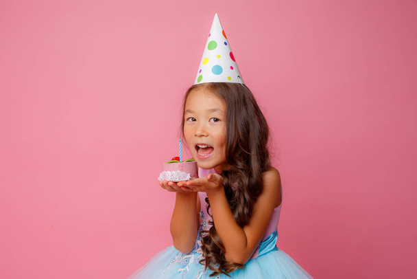 A young Asian girl at a birthday party holds a cake with a candle on a pink background - Фото, изображение