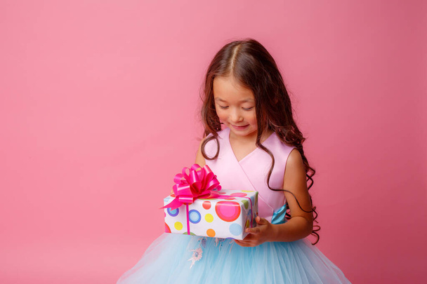 ein kleines Mädchen asiatischen Aussehens hält ein Geschenk in den Händen, das ihren Geburtstag auf rosa Hintergrund feiert - Foto, Bild