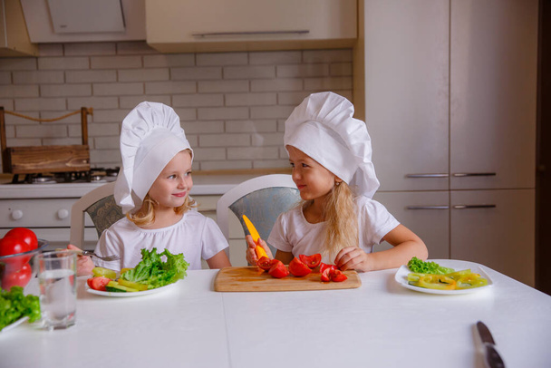 Дві милі смішні дівчата позує в капелюхах шеф-кухаря з овочами на столі
 - Фото, зображення