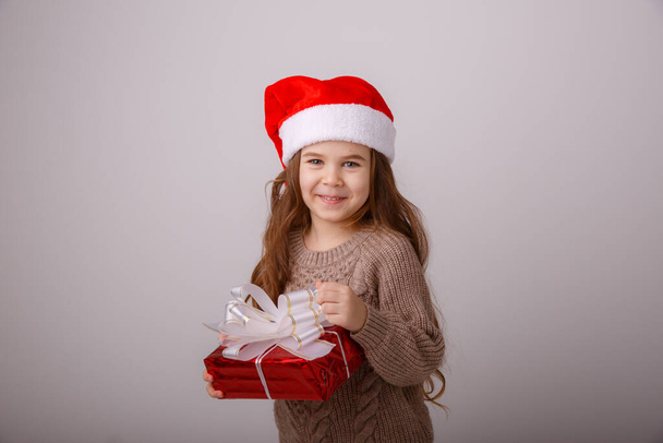 Ένα κοριτσάκι με καπέλο Άι Βασίλη να κρατάει ένα κουτί δώρου ενάντια στο γκρι  - Φωτογραφία, εικόνα