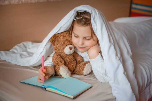 dítě dívka skrývající se pod přikrývkou, která čte knihu s medvídkem  - Fotografie, Obrázek