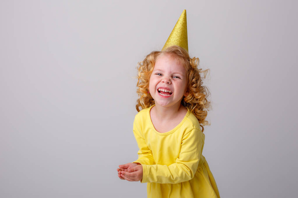 holčička ve žlutých šatech v narozeninové čepici pózující na šedém pozadí - Fotografie, Obrázek