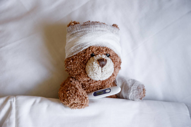 un oso de peluche enfermo está acostado en la cama con un termómetro - Foto, Imagen
