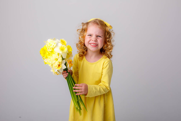 egy kis göndör hajú lány sárga ruhában, egy csokor tavaszi virággal a kezében.   - Fotó, kép