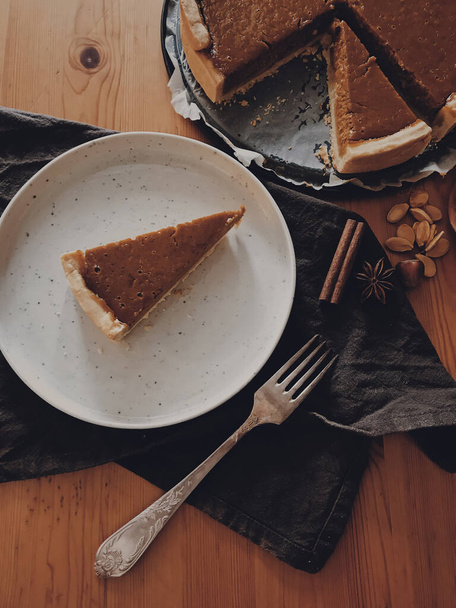 Счастливого Дня Благодарения! Домашний ломтик тыквенного пирога на стильной тарелке со старинной вилкой, анисом и корицей и льняной салфеткой на деревянном столе, вид сверху. Тыквенный пирог на деревенском столе - Фото, изображение