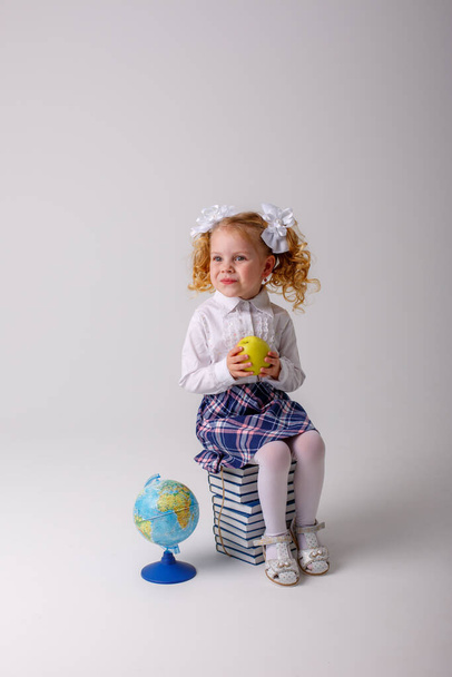 egy kis göndör hajú iskolás lány iskolai egyenruhában, kezében egy almával, egy halom könyvön ülve, - Fotó, kép