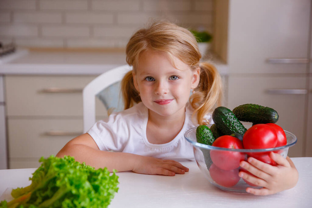 blonde baby girl eating vegetables in the kitchen - Фото, изображение