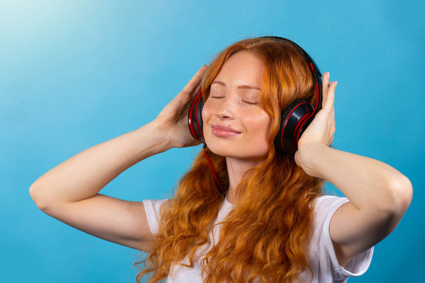 Young beautiful redhaired girl in a white t-shirt enjoying the music with big headphones. Photo on a blue background. - Фото, зображення