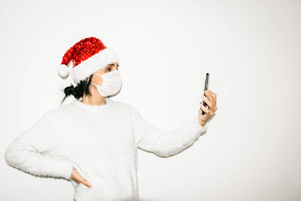 Νεαρή γυναίκα με μάσκα στο πρόσωπο κρατά το κινητό ψηλά απομονωμένο σε λευκό φόντο. Έννοια του προσώπου και της επικοινωνίας κατά τη διάρκεια των διακοπών σε πανδημία. - Φωτογραφία, εικόνα