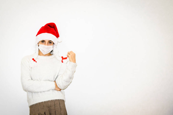 Vrouw in Chrsitmas outfit staat nog steeds bij de witte muur en ziet er recht met lege ruimte aan de rechterkant. Concept van kerstkleding en stemming 2020. - Foto, afbeelding