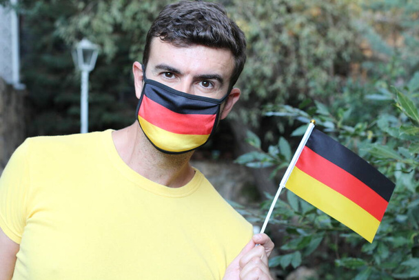 πορτρέτο του όμορφου νεαρού άνδρα στη γερμανική χρωματιστή μάσκα αναπνοής κρατώντας γερμανική σημαία στο πάρκο - Φωτογραφία, εικόνα