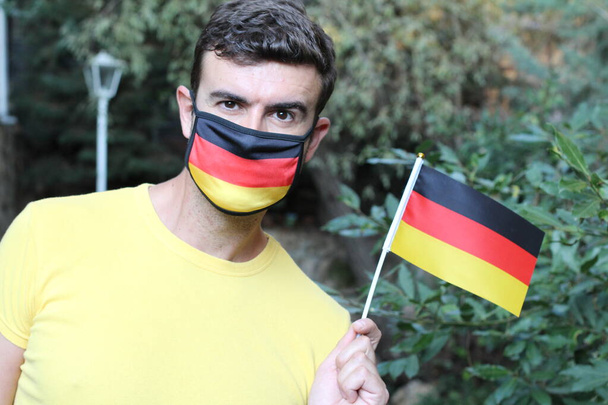 πορτρέτο του όμορφου νεαρού άνδρα στη γερμανική χρωματιστή μάσκα αναπνοής κρατώντας γερμανική σημαία στο πάρκο - Φωτογραφία, εικόνα