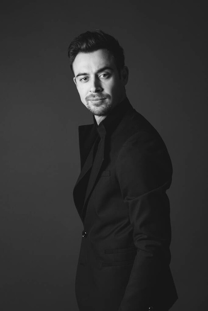 Studio portrait of a young caucasian man in a black blazer, looking at the camera, standing half-turned against plain studio background - Foto, afbeelding