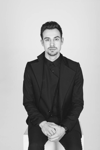 Studio portrait of a young caucasian man in a black jacket, hands together, seriously looking at the camera, sitting against plain studio background - Photo, Image