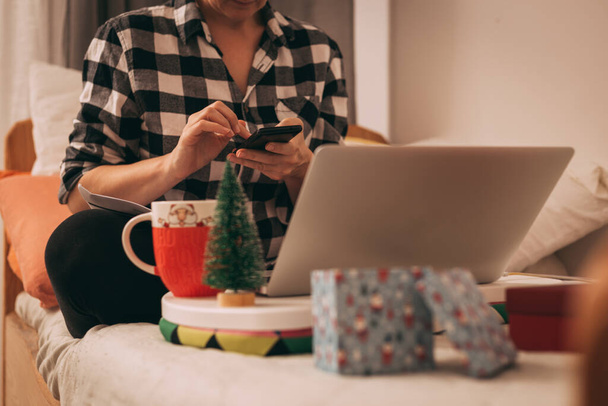 Kobieta w koszuli w kratę za pomocą telefonu komórkowego i laptopa w otoczeniu Bożego Narodzenia - Zdjęcie, obraz