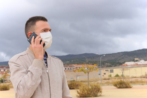 Mann mit wiederverwendbarer Gesichtsmaske benutzt Smartphone im urbanen Hintergrund. Mehrwegmasken sind eine ökologische Alternative zu Einwegmasken wie KN95, FFP2 oder chirurgischen Masken. - Foto, Bild
