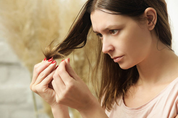 Γυναίκα κοιτάζει στεγνά και μπερδεμένα άκρα των μαλλιών της. - Φωτογραφία, εικόνα