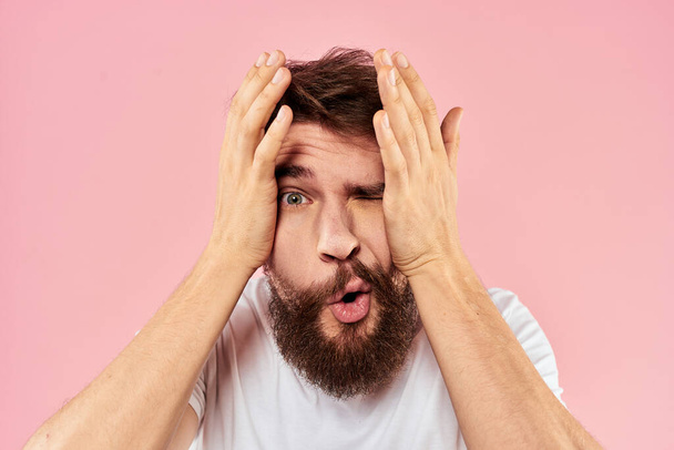 Бородатый мужчина в белой футболке жестикулирует с выражением лица рук вблизи розового фона - Фото, изображение