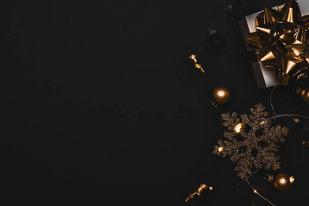 Geschenkband vorhanden. Weiße Geschenkschachtel mit goldenem Band, Neujahrskugeln und funkelnden Lichtergirlanden in weihnachtlicher Komposition auf schwarzem Hintergrund als Grußkarte. Weihnachten, Winter, Neujahrskonzept - Foto, Bild
