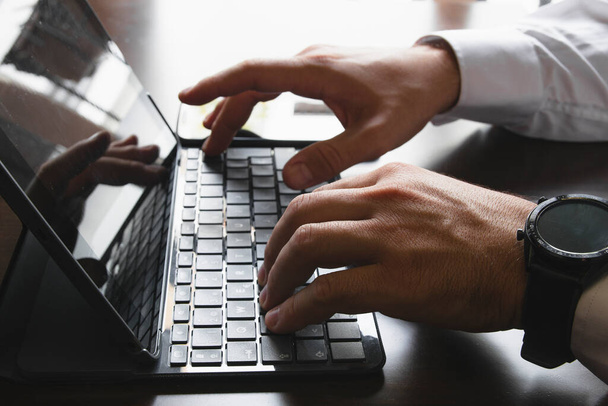 Primo piano delle mani di un uomo in camicia che digita sulla tastiera di un tablet o laptop. Al polso indossa uno smartwatch e uno smarhphone sullo sfondo. Fotografia orizzontale - Foto, immagini