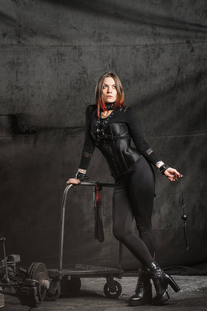 Femme dominante sexy avec des piercings et des cheveux brillants dans un corset noir, avec harnais et bracelets en cuir, tient un bâillon, jouets pour adultes, regarde strictement, sur un fond industriel - Photo, image