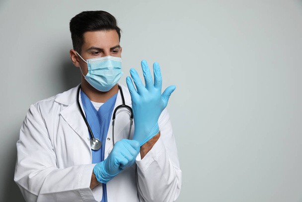 Doctor in protective mask putting on medical gloves against light grey background - Foto, Bild