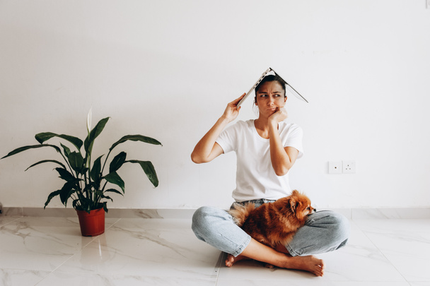 Female portrait with laptop above head. Showing expression girl sitting in white t-shirt on the floor with pomeranian dog. Woman holding laptop. Quarantine. Stay at home. - Photo, image